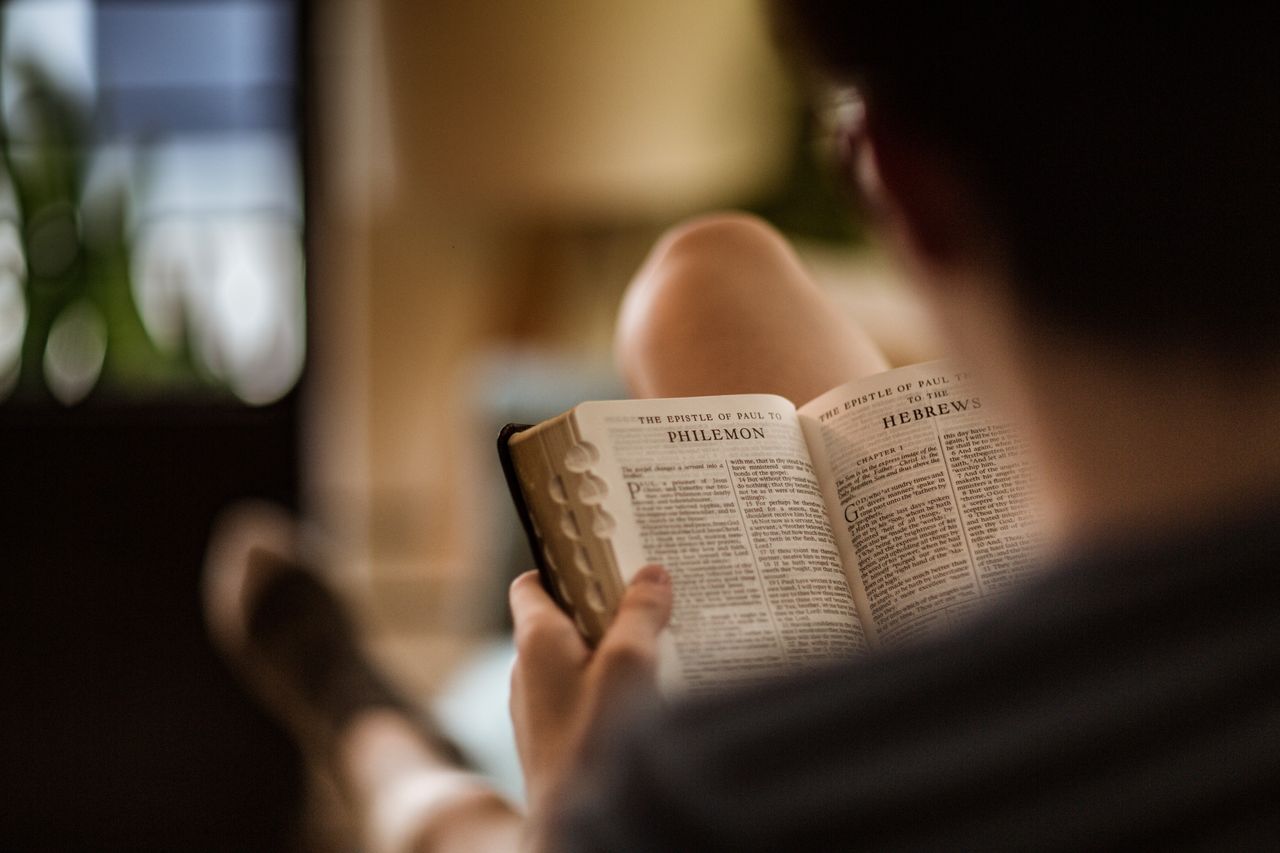 Muž sedí na pohovce a studuje Bibli svatou, která je otevřená na stránkách s epištolou, kterou Pavel napsal Filemonovi